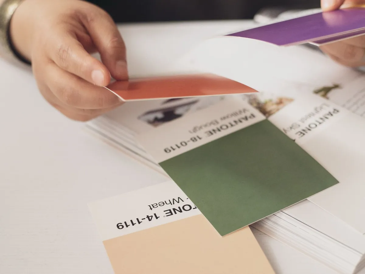 Manos que eligen muestras de color impresas sobre una mesa blanca: tarjetas verde, terracota, violeta y beige