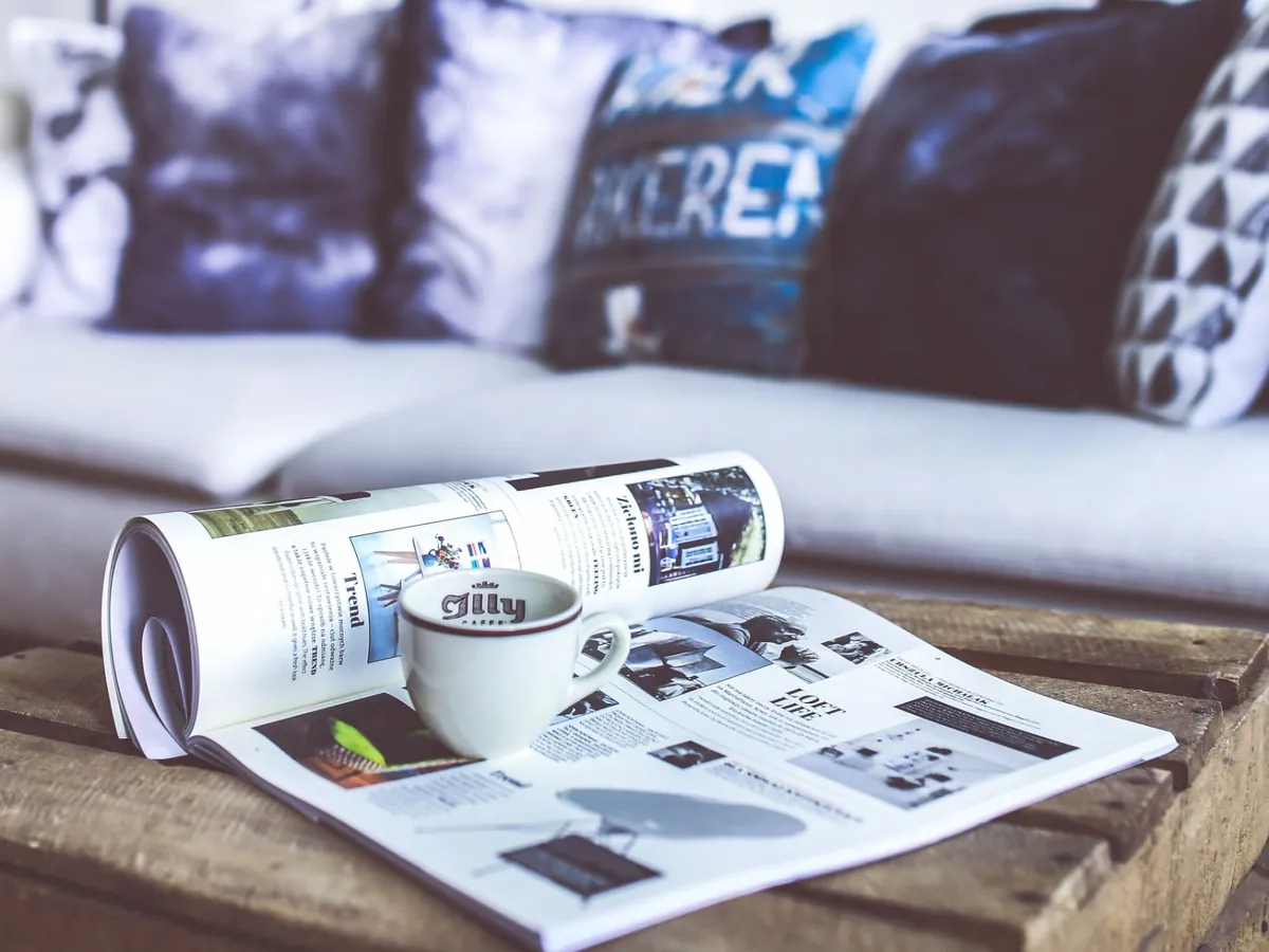 Revista de diseño abierta y una taza sobre una mesa de cajón de madera, frente a un sillón claro con almohadones azules
