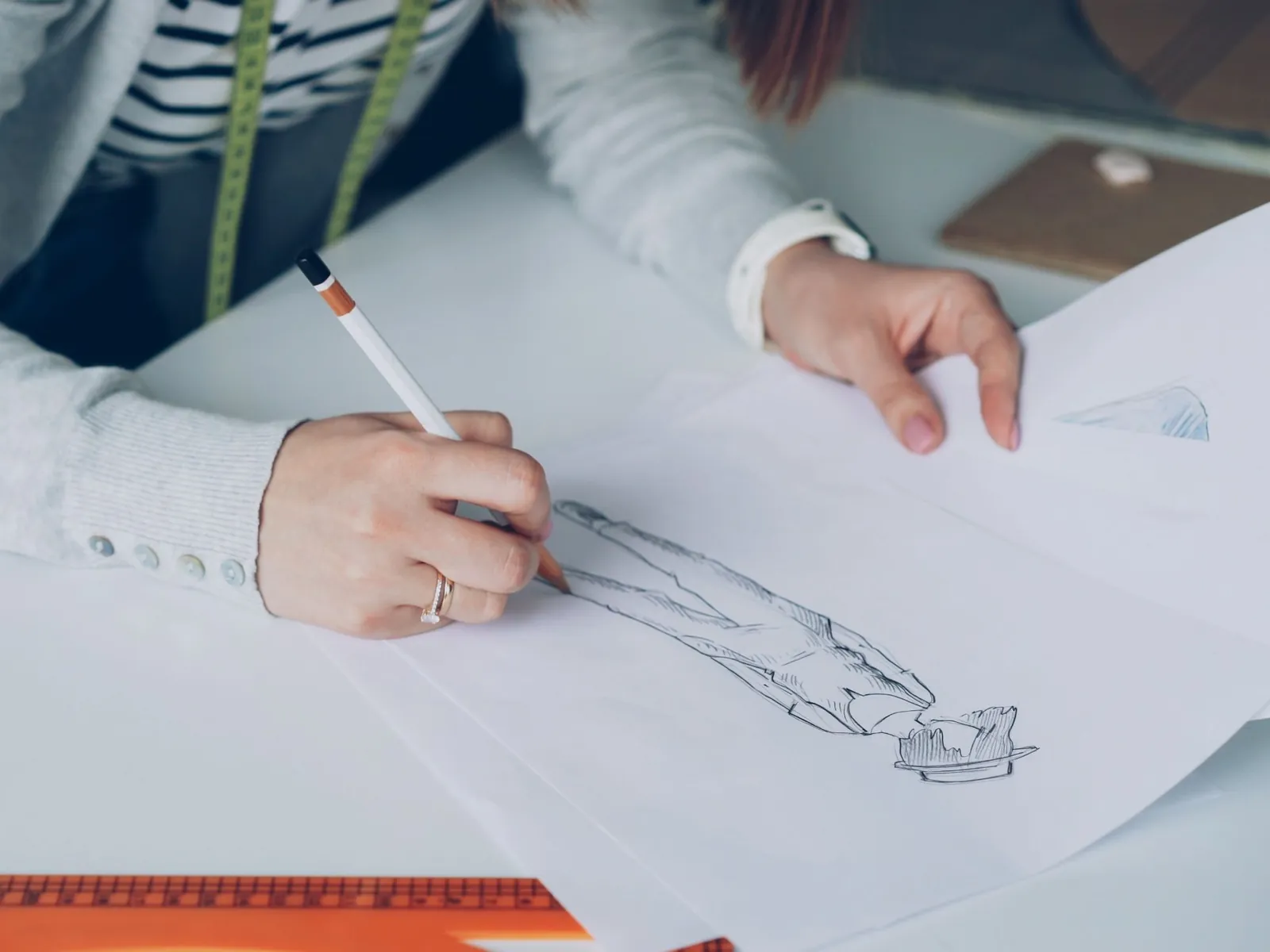 Una diseñadora con cinta métrica al cuello dibuja un figurín a lápiz sobre la mesa de trabajo